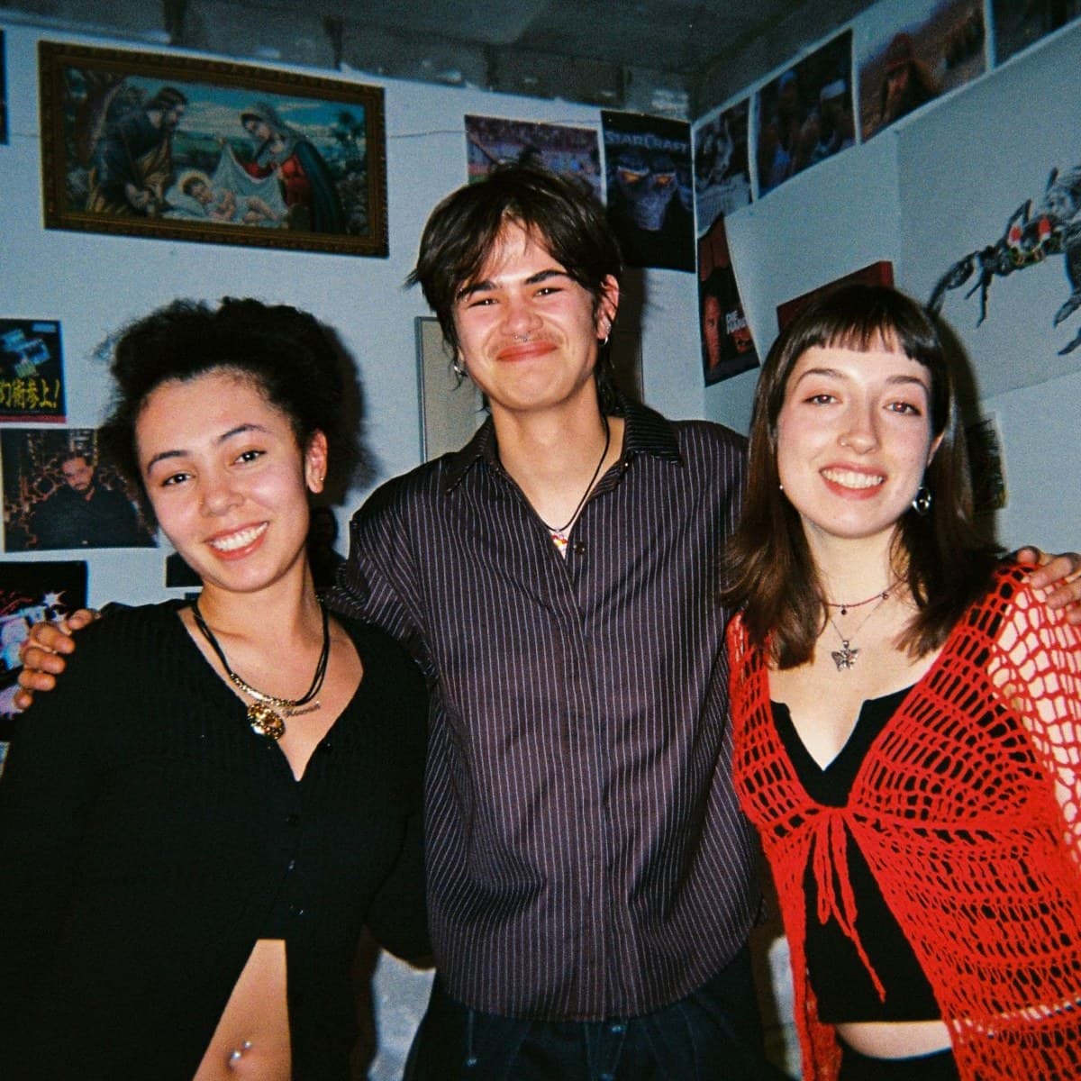 Three artists standing arm in arm and smiling, in a room with photographs, posters and prints covering the wall behind them.