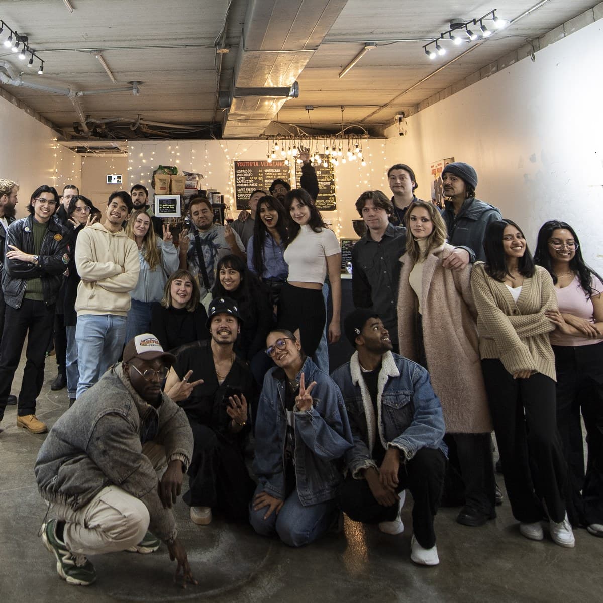 About twenty-five people gathered together for a group photo at a Vengeance Foundation exhibition, artwork pinned to the walls and string lights strung across the ceiling behind them.