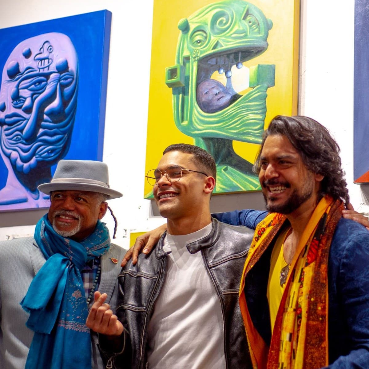 Three people standing together and laughing at a Vengeance Foundation exhibition, in front of large painted portraits hung on the gallery wall.