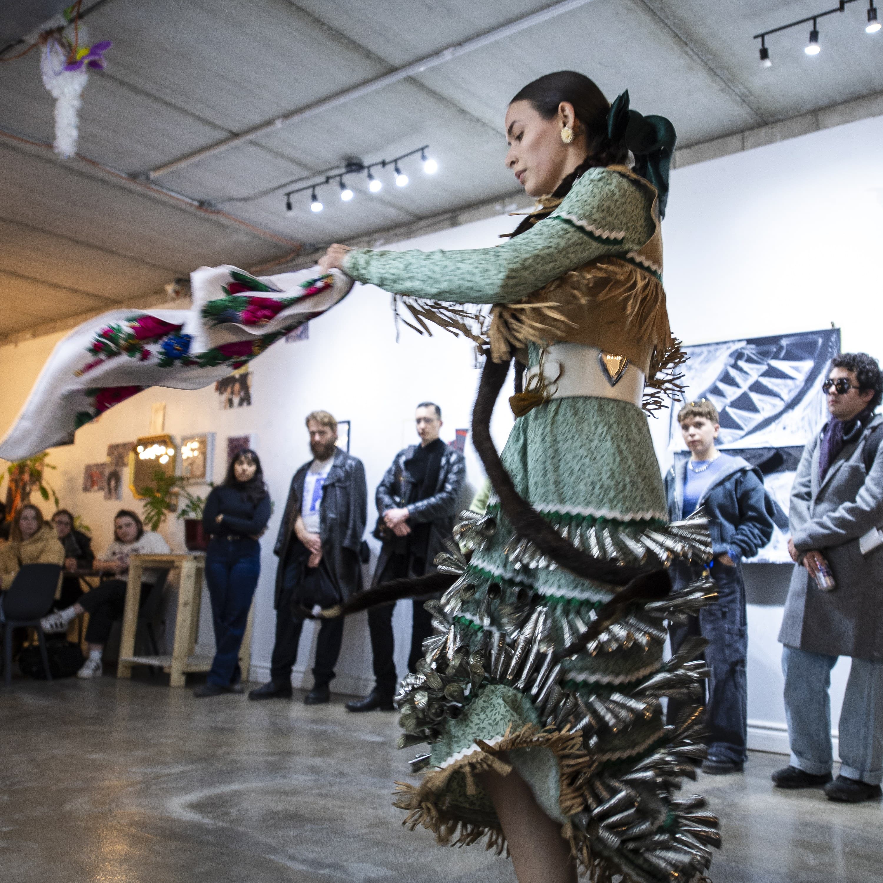 A jingle dress dancer mid-turn with her shawl outstretched, performing for a standing and seated audience in a gallery space.