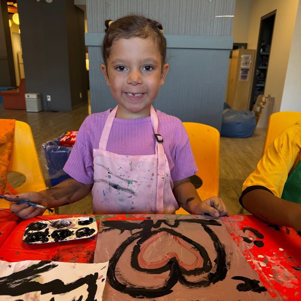 A young child in a paint-splattered apron smiling behind the heart she has just painted, at a Vengeance in Action kids workshop.